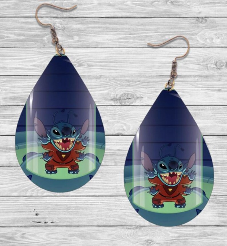 May include: A pair of teardrop-shaped earrings featuring a blue and green image of Stitch from Lilo & Stitch. Stitch is in a glass tube wearing a red and blue spacesuit. The earrings have a silver hook.