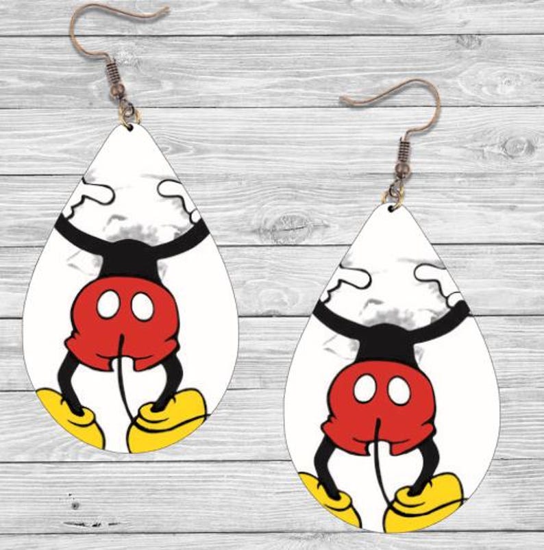 Op de afbeelding: Een paar druppelvormige oorbellen met een cartoonkarakterontwerp. De oorbellen hebben een witte achtergrond met een zwarte, rode en gele karakterillustratie. De oorbellen hangen aan een koperkleurige haak.