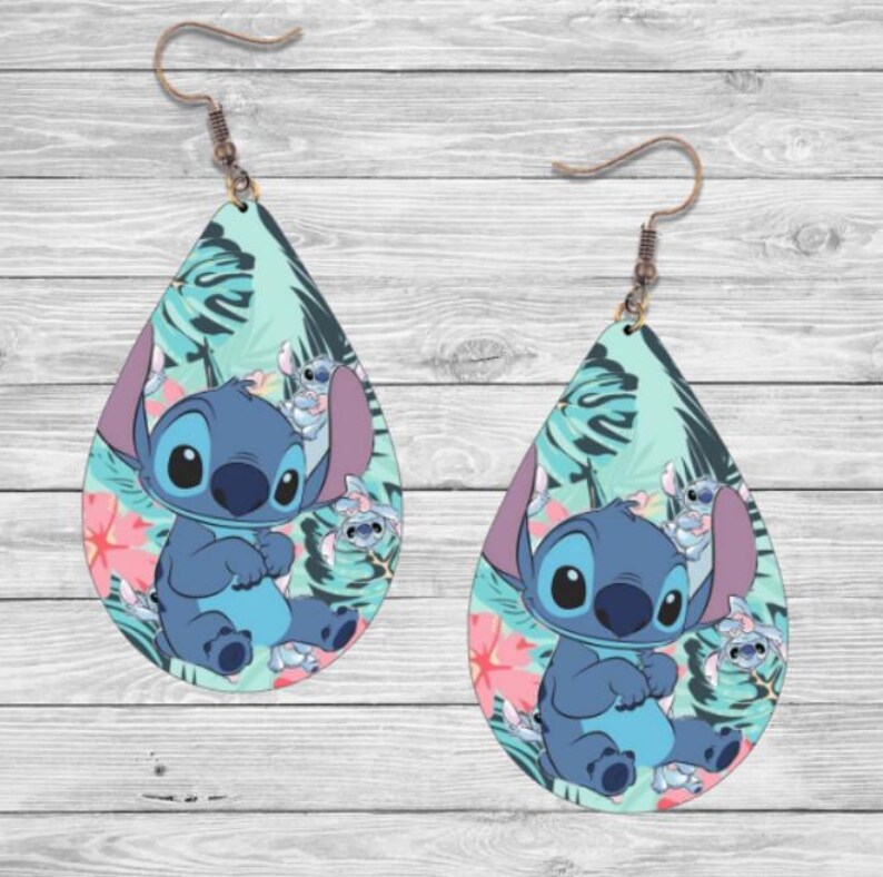 May include: A pair of teardrop-shaped earrings featuring a blue cartoon character with large eyes and a wide smile. The character is surrounded by tropical leaves and flowers in shades of green, pink, and blue.