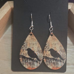 K&ouml;nnte beinhalten: Ein Paar tropfenf&ouml;rmige Ohrringe mit Vintage-Design. Sie zeigen eine schwarze Vogelsilhouette auf einem Ast vor einem Hintergrund aus gealtertem Papier mit orangefarbenen und braunen Akzenten. Die Ohrringe h&auml;ngen an silberfarbenen Haken.