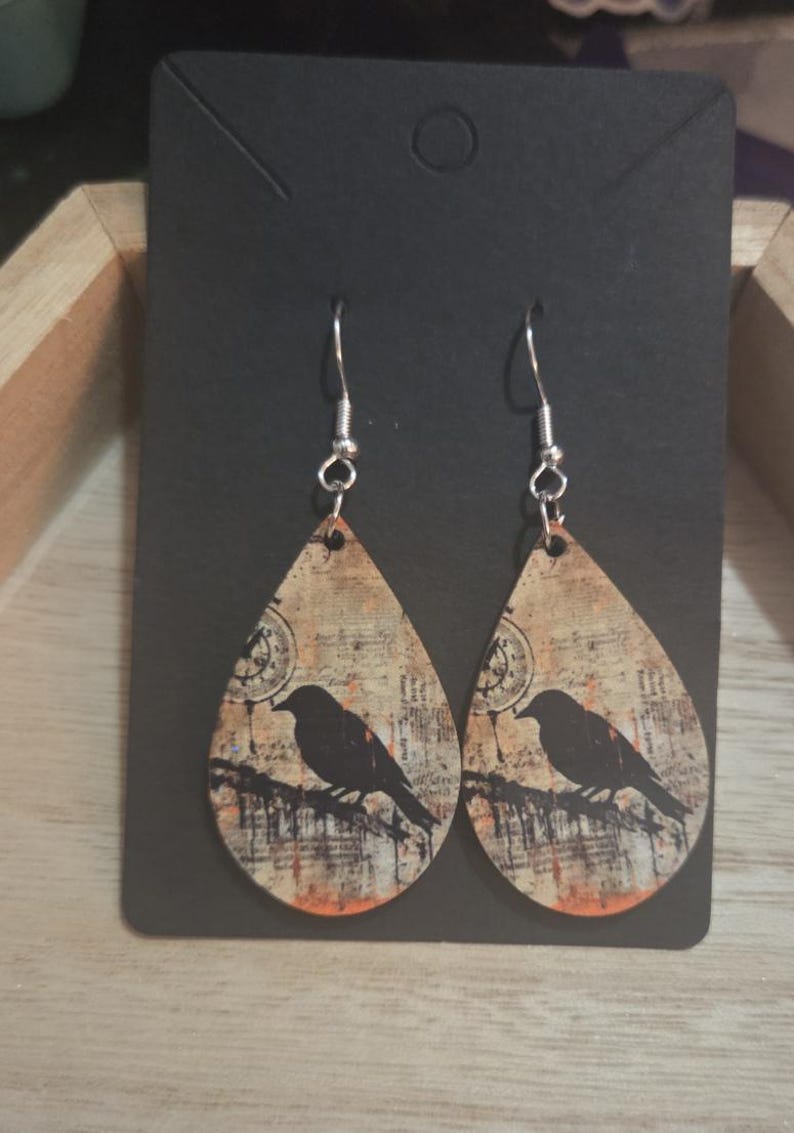 K&ouml;nnte beinhalten: Ein Paar tropfenf&ouml;rmige Ohrringe mit Vintage-Design. Sie zeigen eine schwarze Vogelsilhouette auf einem Ast vor einem Hintergrund aus gealtertem Papier mit orangefarbenen und braunen Akzenten. Die Ohrringe h&auml;ngen an silberfarbenen Haken.