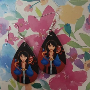 May include: A pair of teardrop-shaped dangle earrings featuring a printed illustration of Mulan holding a sword with Mushu the dragon wrapped around her shoulders. The earrings have silver-toned metal fishhook wires for pierced ears.