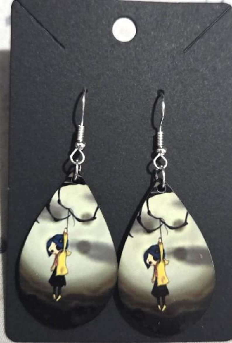 Peut inclure: Boucles d'oreilles en forme de goutte d'eau avec une illustration de dessin anim&eacute; d'une fille en manteau jaune suspendue &agrave; une branche sur fond de ciel nuageux. Les boucles d'oreilles ont des crochets d'oreille argent&eacute;s.