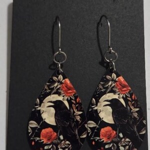 K&ouml;nnte beinhalten: Tropfenf&ouml;rmige Ohrringe mit schwarzem Hintergrund, die einen Raben vor einem Vollmond zeigen, umgeben von roten Rosen und Bl&auml;ttern. Die Ohrringe h&auml;ngen an silberfarbenen Haken.