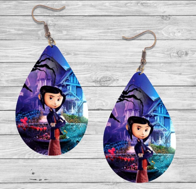 Peut inclure: Boucles d'oreilles en forme de goutte d'eau avec un personnage de dessin anim&eacute; devant une maison. Les boucles d'oreilles ont un fond bleu fonc&eacute; avec un motif de maison et d'arbre. Le personnage porte une veste noire et une jupe marron.