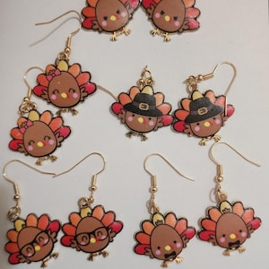 May include: A collection of gold-toned dangle earrings featuring cartoon turkey designs. The turkeys have brown bodies, colorful tail feathers, and various accessories like hats and glasses. The earrings are likely for the Thanksgiving holiday.