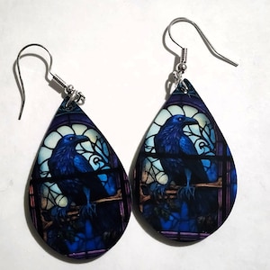 K&ouml;nnte beinhalten: Ein Paar tropfenf&ouml;rmige Ohrringe mit einem Buntglasdesign, das einen blauen Raben auf einem Ast zeigt. Die Ohrringe haben silberfarbene Fischhaken-Ohrdr&auml;hte und ein dunkelblaues und violettes Farbschema.