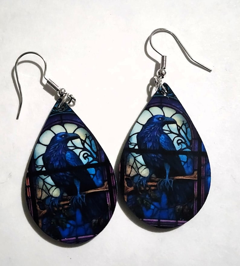 K&ouml;nnte beinhalten: Ein Paar tropfenf&ouml;rmige Ohrringe mit einem Buntglasdesign, das einen blauen Raben auf einem Ast zeigt. Die Ohrringe haben silberfarbene Fischhaken-Ohrdr&auml;hte und ein dunkelblaues und violettes Farbschema.