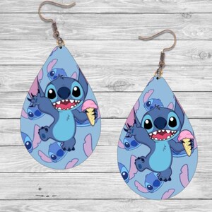 May include: A pair of blue teardrop-shaped earrings featuring a cartoon character holding an ice cream cone. The character is blue with large eyes and a wide smile. The background is a light blue with a pattern of the same character.