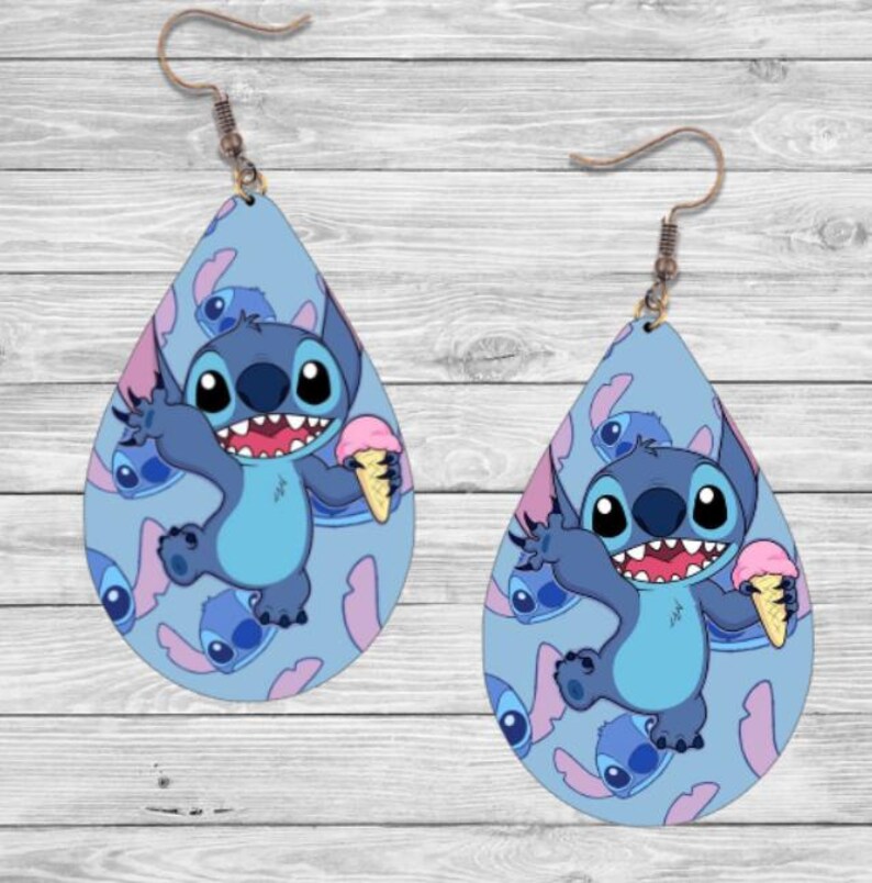 May include: A pair of blue teardrop-shaped earrings featuring a cartoon character holding an ice cream cone. The character is blue with large eyes and a wide smile. The background is a light blue with a pattern of the same character.