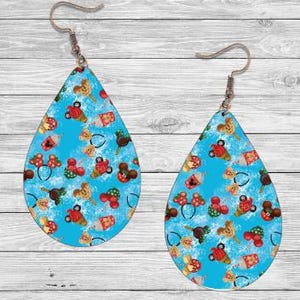 Op de afbeelding: Druppelvormige oorbellen met een lichtblauwe achtergrond en een herhalend patroon van stripfiguren in rood, bruin en geel. De oorbellen hebben zilverkleurige oorhaken.