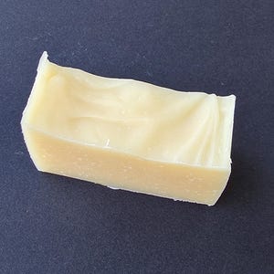 Puede incluir: Una pastilla rectangular de jabón artesanal de color amarillo claro. El jabón tiene una superficie ligeramente texturizada y una parte superior ondulada. El jabón está sobre un fondo azul oscuro.