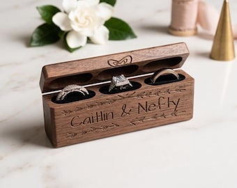 Könnte beinhalten: Eine rechteckige Ringbox aus dunklem Walnussholz mit Klappdeckel und drei runden, schwarz mit Samt ausgekleideten Fächern für drei Ringe. Die Vorderseite ist mit dem Text "Caitlin & Nefty" und Pfeilmotiven graviert, auf dem Deckel ist ein Herz.