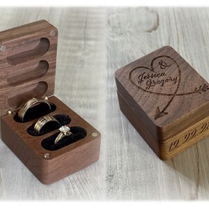Puede incluir: Una caja de anillos de madera con un diseño de corazón y flecha grabado en la tapa. La tapa está abierta para revelar tres ranuras para anillos forradas con terciopelo negro. La caja está grabada con el texto "Jessica & Gregory" y la fecha "12.22.23".