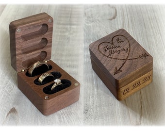 May include: A wooden ring box with a heart and arrow design engraved on the lid. The lid is open to reveal three ring slots lined with black velvet. The box is engraved with the text "Jessica & Gregory" and the date "12.22.23".