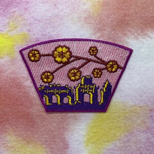 May include: Purple and gold embroidered patch with a cityscape and a branch of flowers with yellow blossoms. The patch is shaped like a half-circle.