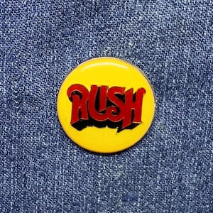 May include: A circular pinback button featuring a bright yellow background with the word RUSH written in bold red stylized lettering with a black drop shadow effect. The pin is centered against a textured blue denim fabric background.