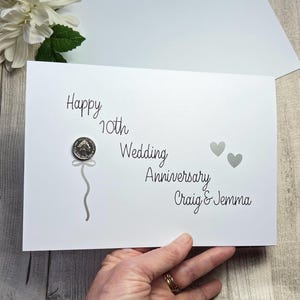 May include: A white anniversary card with the text "Happy 10th Wedding Anniversary Craig & Jemma" in black script. A silver coin is attached to the card with a grey ribbon design. Two grey hearts are also on the card.