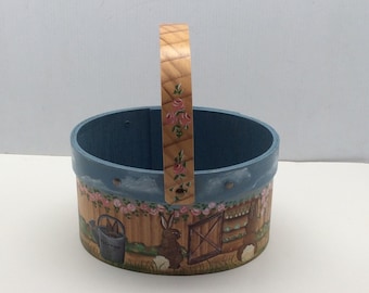 May include: A blue and brown painted wooden Easter basket with a handle. The basket is decorated with a floral design and a scene of rabbits and a garden.
