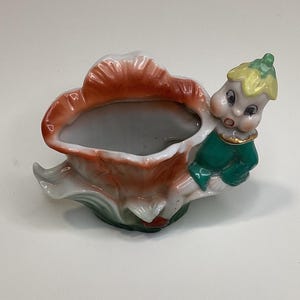Puede incluir: Un macetero de cerámica con forma de flor con una pequeña figura de duende sujetando el lateral. El macetero es blanco con detalles en naranja y verde. El duende lleva una camisa verde y un gorro amarillo.