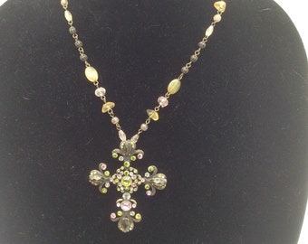 May include: A gold necklace with a large cross pendant. The cross is decorated with colorful rhinestones and has a detailed design. The necklace is made of small beads in shades of yellow, pink, and brown.