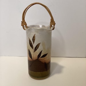 May include: A cylindrical ceramic vase with a woven handle. The vase features a cream-colored top with a brown and tan painted design of leaves and a dark brown base. The handle is made of woven tan material.