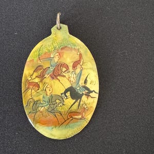 May include: An oval-shaped pendant with a hand-painted scene of three people on horseback riding through a landscape. The pendant is made of wood and has a silver-colored loop at the top for hanging.