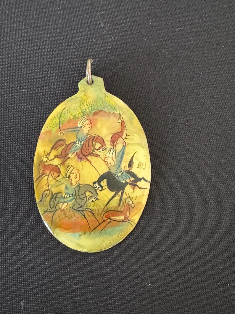 May include: An oval-shaped pendant with a hand-painted scene of three people on horseback riding through a landscape. The pendant is made of wood and has a silver-colored loop at the top for hanging.