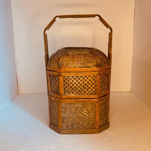 May include: A brown, multi-tiered, hexagonal woven basket with a sturdy arched handle. The structure features two stacked compartments with intricate lattice and diagonal weave patterns, topped with a matching woven lid and a vertical frame.
