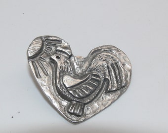 May include: A silver-colored heart-shaped brooch with a detailed relief design. The brooch features a rooster and a sun, with textured details throughout. The design is raised, creating a three-dimensional effect.
