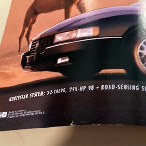 May include: A black Cadillac Sedan DeVille with a Northstar System 32-valve, 295-hp V8 engine and road-sensing suspension. The car is driving on a desert road with a camel in the background.