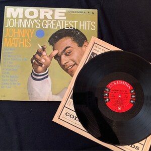 May include: A vintage vinyl record album cover for "More Johnny's Greatest Hits" by Johnny Mathis. The album cover is yellow and features a photo of Johnny Mathis. A black vinyl record is next to the album cover.