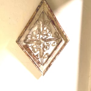 Peut inclure: Décoration murale en forme de losange avec un cadre en bois blanc cassé vieilli et un motif décoratif en filigrane métallique. Le travail du métal présente des motifs tourbillonnants et un médaillon central. Esthétique rustique et vintage.