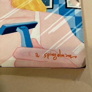 May include: A close-up of a blue razor with a white handle, held by a hand in front of a mirror. The mirror reflects a woman with blonde hair and a pink shirt. The signature "a. spiegelman" is visible in the bottom right corner.
