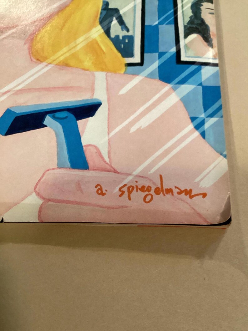 May include: A close-up of a blue razor with a white handle, held by a hand in front of a mirror. The mirror reflects a woman with blonde hair and a pink shirt. The signature "a. spiegelman" is visible in the bottom right corner.