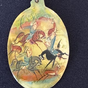 May include: An oval-shaped pendant with a hand-painted scene of three figures on horseback riding through a landscape. The figures are painted in shades of blue, red, and yellow, and the background is a mix of green and brown.