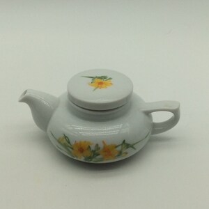 May include: A small white teapot with a floral design. The teapot has a lid with a single yellow flower on it. The teapot is decorated with yellow flowers and green leaves.