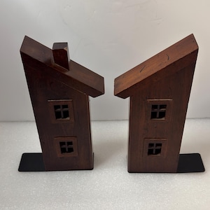 May include: Two wooden house-shaped bookends. The bookends are dark brown with two square windows and a chimney. Each bookend has a black metal base for stability. The bookends are designed to hold books upright.