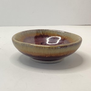 May include: A small, handmade ceramic bowl with a unique color gradient. The interior is a deep burgundy, transitioning to shades of orange and tan towards the rim. The bowl sits on a white base, showcasing the craftsmanship and artistic design.