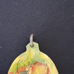 May include: A hand-painted oval pendant with a scene of a man on horseback hunting a deer. The pendant is made of a light brown material and has a silver loop at the top.