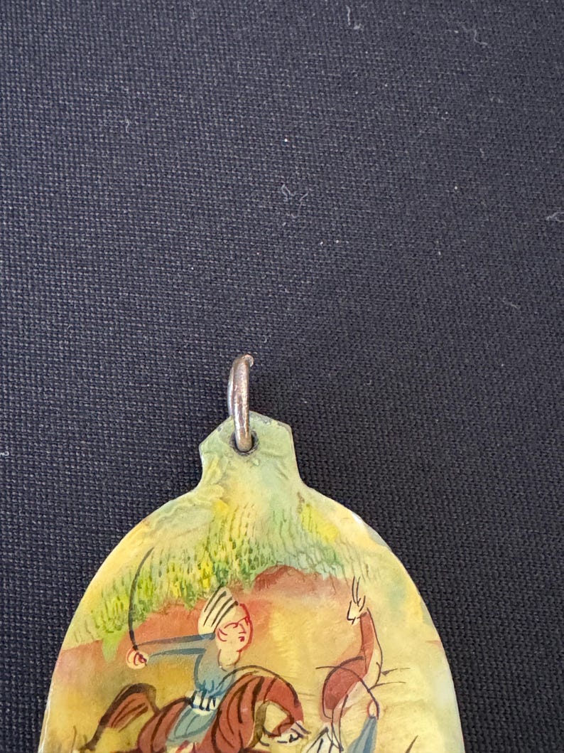 May include: A hand-painted oval pendant with a scene of a man on horseback hunting a deer. The pendant is made of a light brown material and has a silver loop at the top.