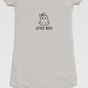 Puede incluir: Body de bebé blanco con un gráfico negro de un fantasma con un lazo y el texto "LITTLE BOO". El body tiene mangas cortas y corchetes en la parte inferior. Una prenda de bebé con temática de Halloween.