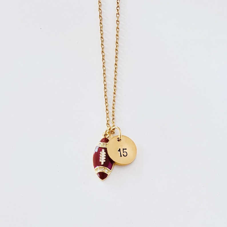 May include: A gold chain necklace with a burgundy football charm and a gold disc charm with the number 15.