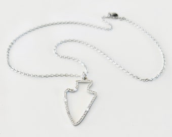 Puede incluir: Un collar de plata con un colgante de punta de flecha de plata. La punta de flecha está bordeada de pequeñas piedras transparentes.