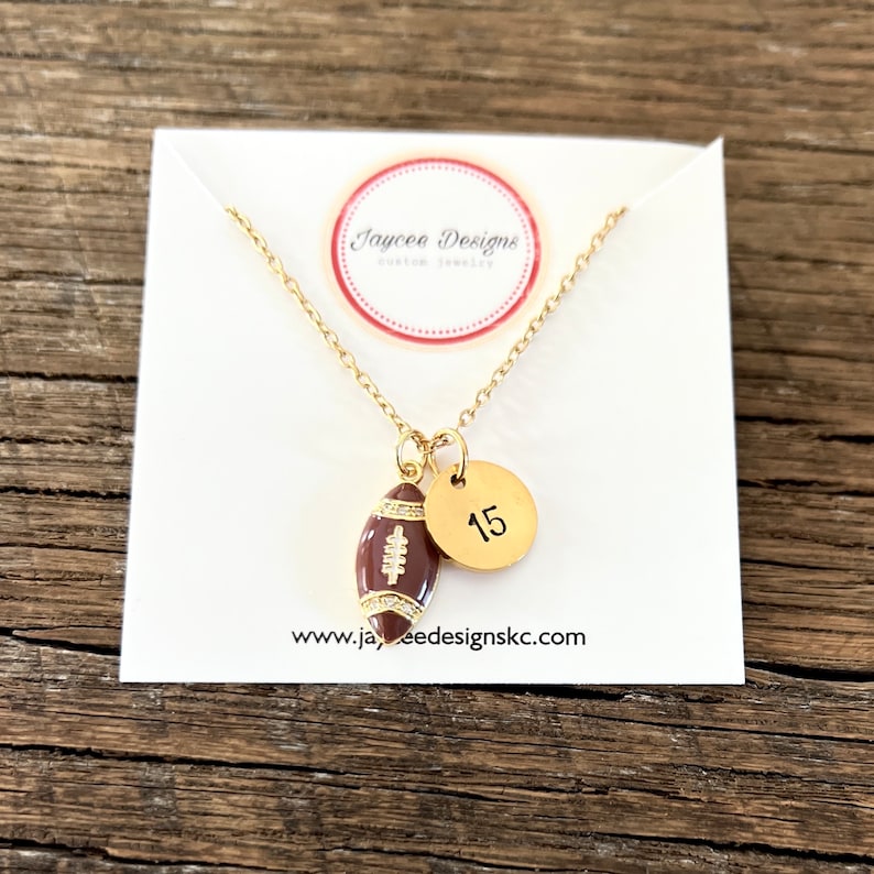 May include: Gold necklace with a brown football charm and a gold circle charm with the number 15 on it. The necklace is on a white card with the text "Jaycee Designs Custom Jewelry" and "www.jayceedesignskc.com".