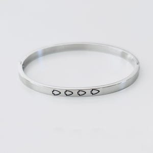 May include: A silver-colored bangle bracelet with a flat surface. The bracelet features a series of four black arrow symbols engraved along the front. The bracelet is circular and appears to be made of metal.
