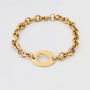 May include: A gold-toned chain link bracelet featuring a central circular charm with a cutout in the shape of an arrowhead. The bracelet is constructed from uniform round links and includes a secure lobster claw clasp closure at the top.
