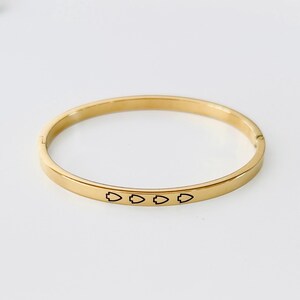 May include: Gold-colored bangle bracelet with a flat, rectangular design. The bracelet features a series of small, black arrow-shaped symbols engraved along the front. The bracelet is shown on a white background.