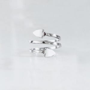 May include: A silver-toned metal ring featuring a bypass design with two pointed arrow-shaped ends. The band wraps around in a spiral shape, creating an open-ended structure with a polished, reflective surface finish throughout the entire piece.