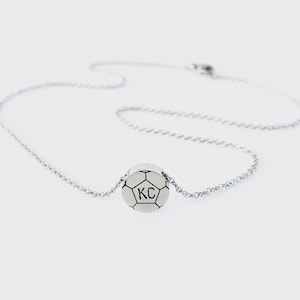 May include: A silver chain necklace with a small silver soccer ball pendant. The soccer ball has the initials "KC" engraved on it.